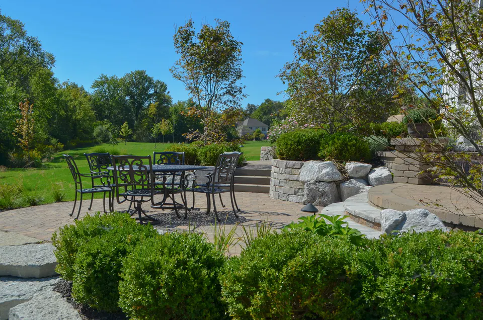 Paver patio seating area with stone retaining wall, steps, shrubs, and open lawn beyond.